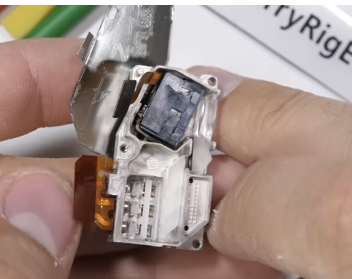 Close-up of the Blackview Max 1's laser-projector optical engine held between two fingers: the black MEMS scanning micro-mirror module at the top, and the three RGB laser diodes with a beam-combining crystal on the lower stage.